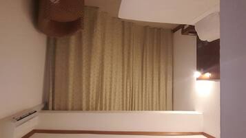 A hotel room with a large window, a curtain, a wall-mounted air conditioning unit, and a wooden trim along the baseboard.
