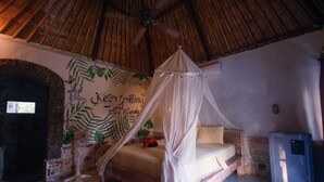 Una habitación con cama bajo dosel, un mural en la pared, ventilador de techo y refrigerador.