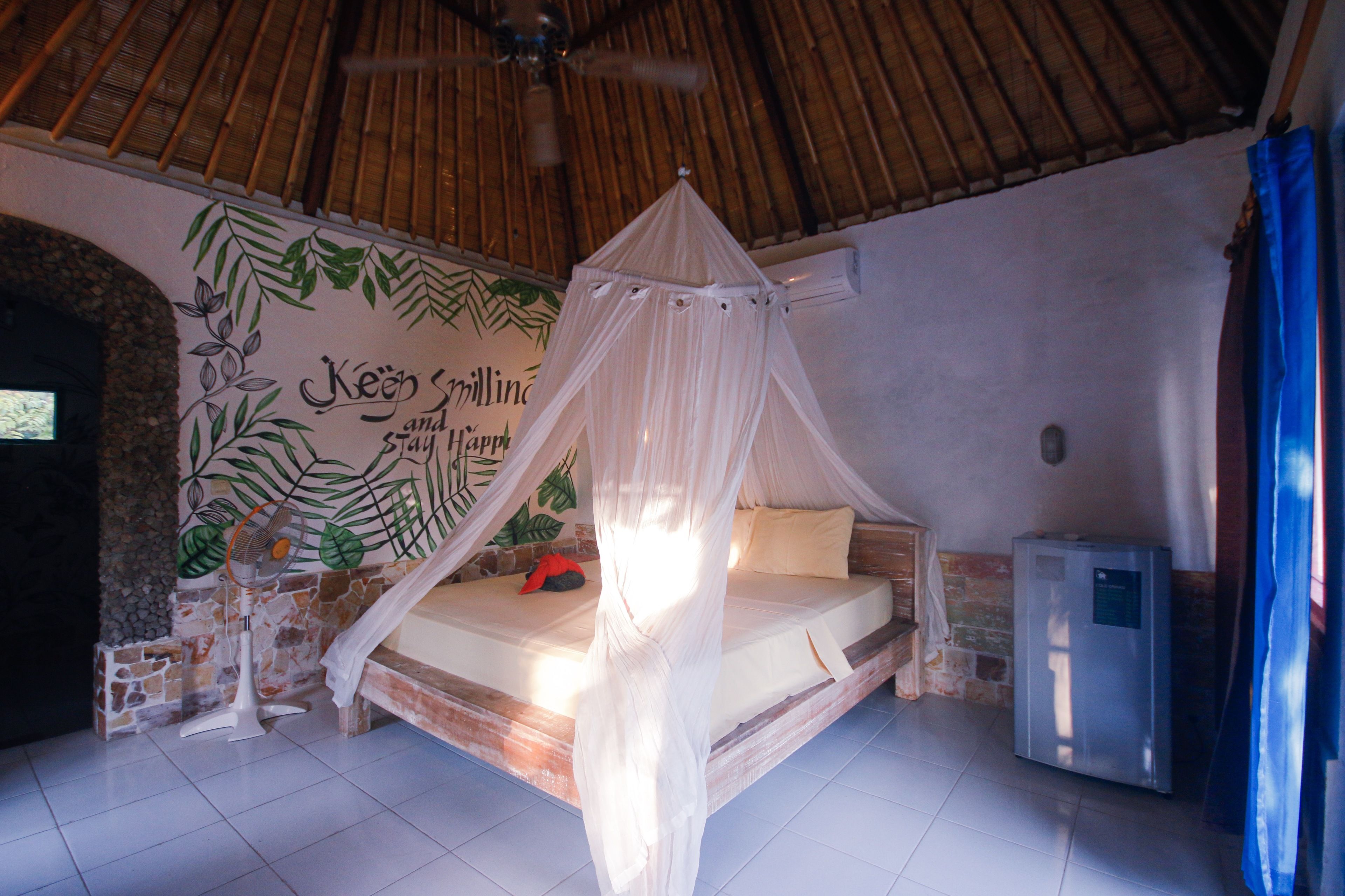 A room with a bed under a canopy, a wall mural, a fan, and a small refrigerator.