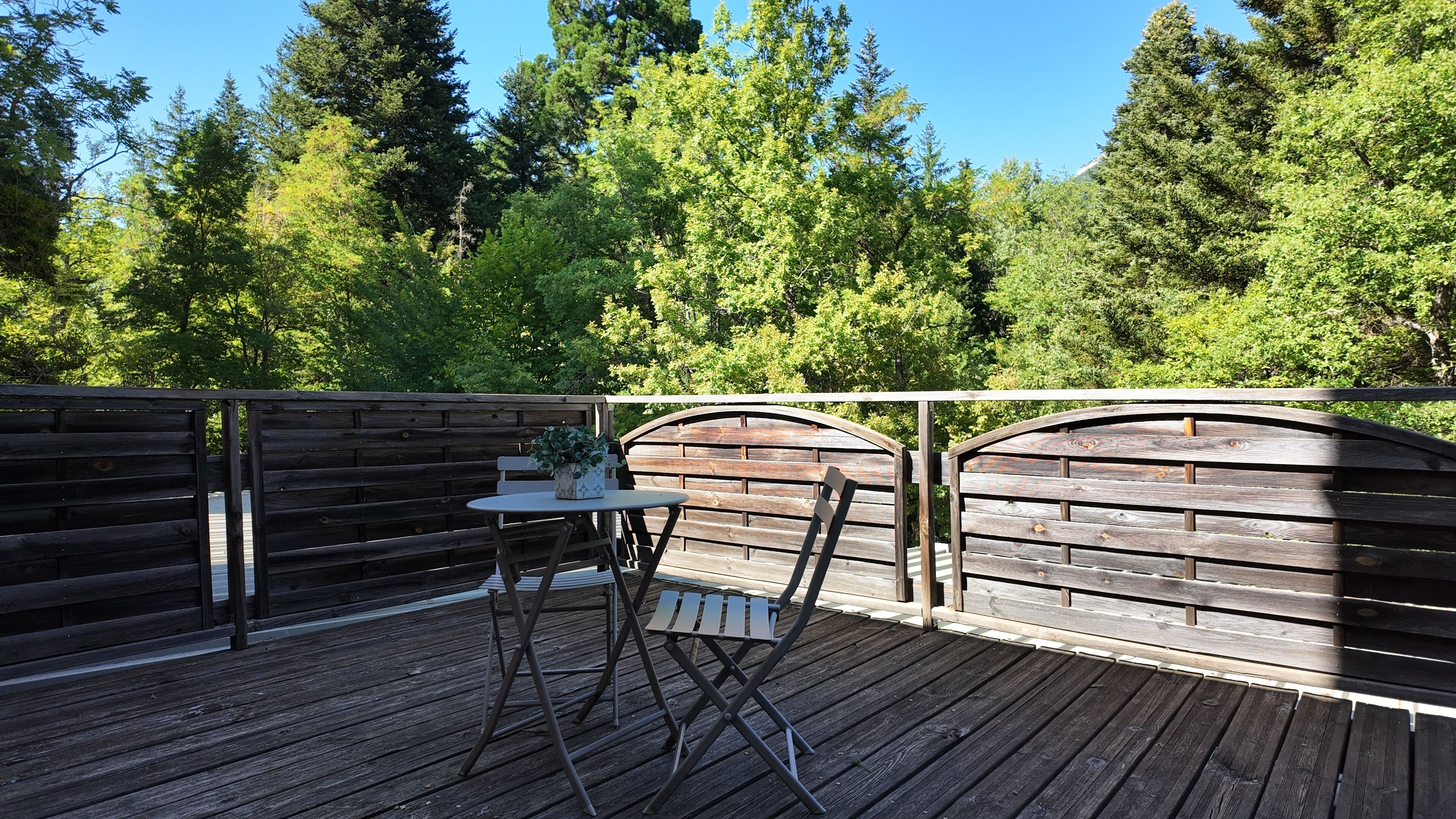 A wooden deck with a table and chairs, surrounded by a wooden fence and dense green trees.