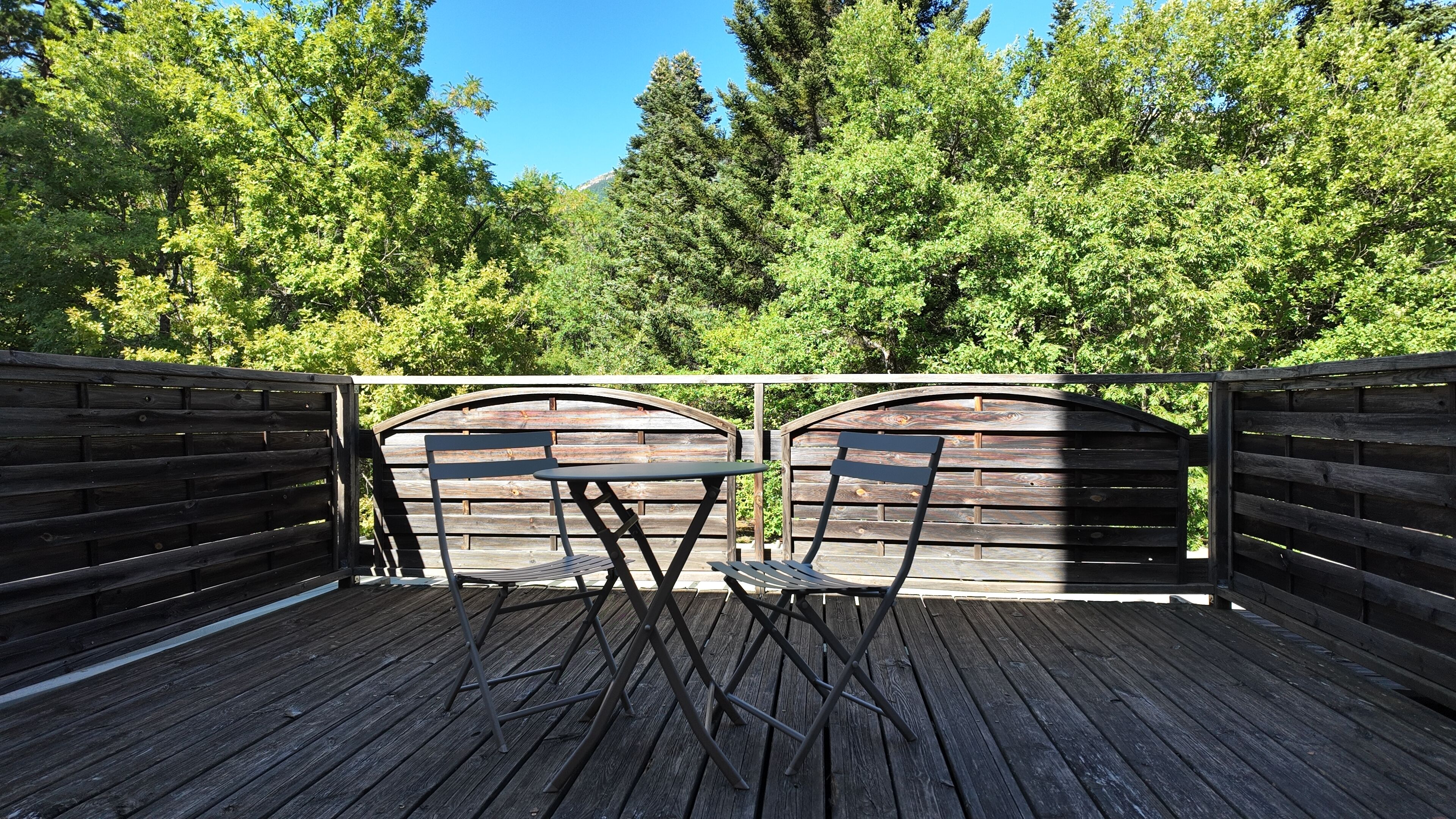 An outdoor wooden deck with two tables and chairs, surrounded by a wooden fence and dense green foliage.