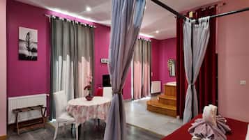 A room with a pink wall, a round table with a floral tablecloth, a white chair, a wooden chair, a small wooden stool, a red carpet, and a white towel on a red mat.