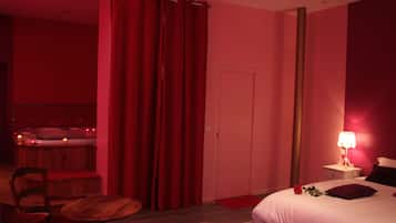Une chambre d’hôtel avec une chaise en bois, une petite table ronde, un lit avec un oreiller et une rose, une table de chevet avec une lampe, et un rideau rouge.