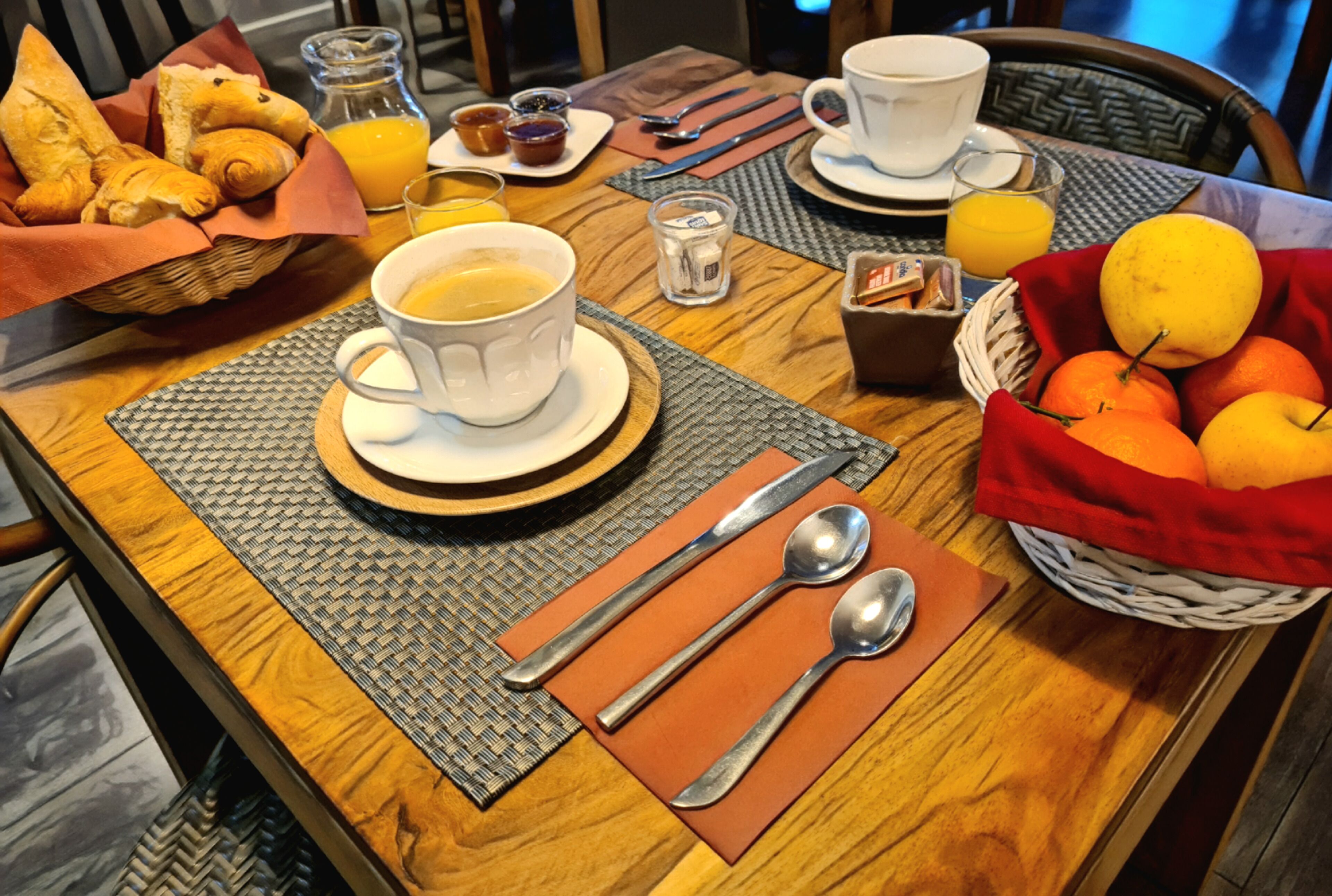 A breakfast table with a cup of coffee, a glass of orange juice, a basket of oranges, and a wicker basket with croissants.