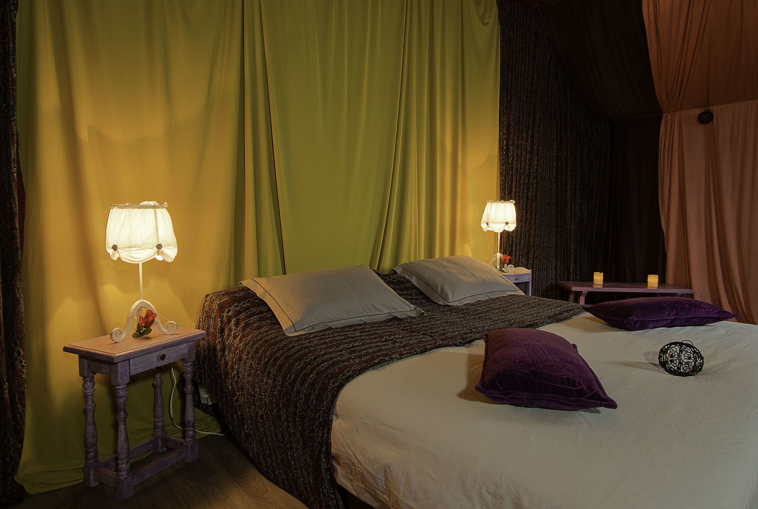 Een keurig opgemaakt bed met kussens, een nachtkastje met een lamp en een gordijnenachtergrond.