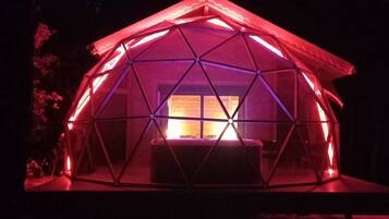 A geodesic dome structure with a central window, illuminated by red lighting, set in a forested area.