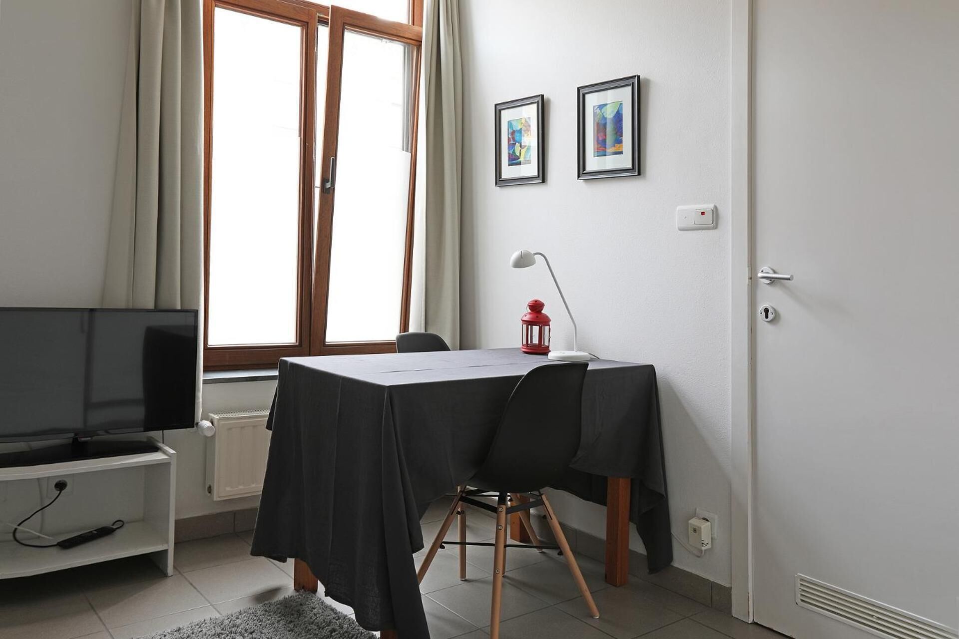 Une petite pièce bien éclairée, équipée d’une télévision, d’une table avec une chaise, d’une fenêtre avec des rideaux et de deux tableaux encadrés accrochés au mur.