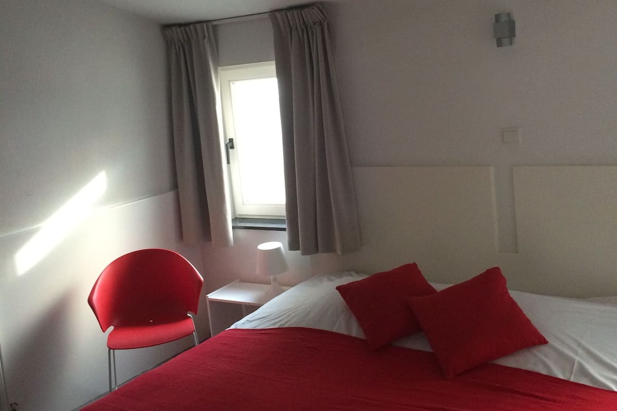 A hotel room with a bed, red pillows, a red chair, a small table, and a window with curtains.
