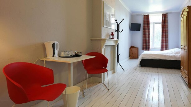 Une chambre comprenant un lit, un bureau avec une bouilloire, deux chaises rouges, un porte-manteau et un téléviseur.