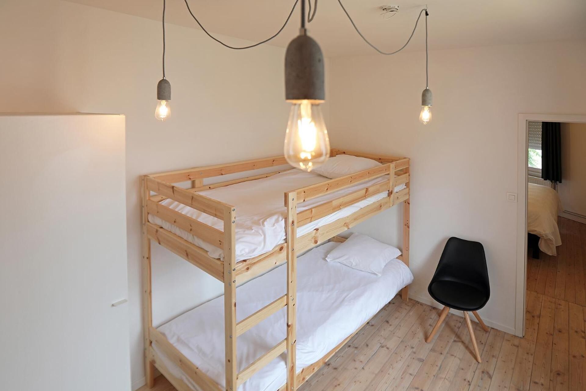 A bunk bed with a wooden frame and white bedding, a black chair, and a hanging light fixture with exposed bulbs.