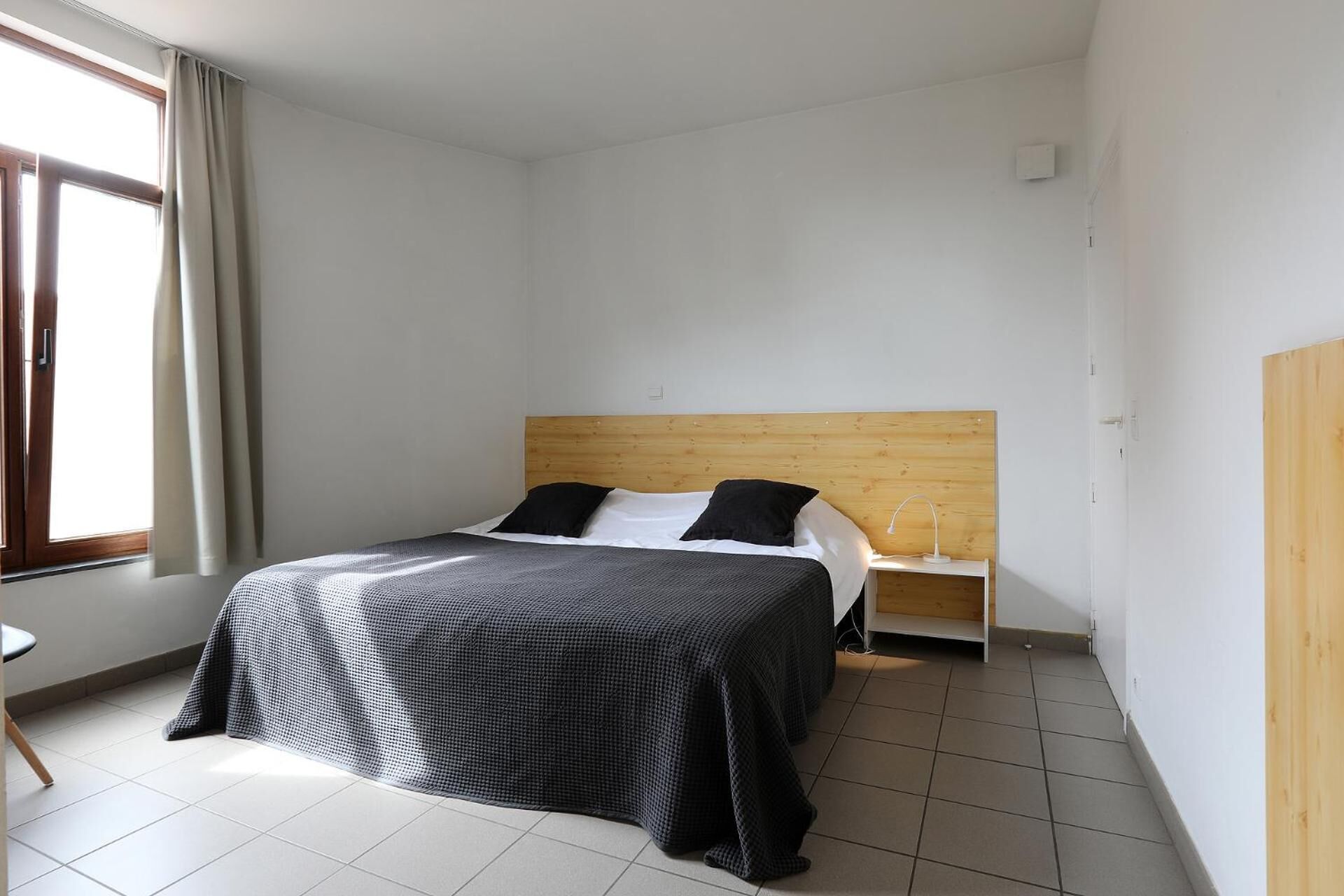 A hotel room with a bed, a nightstand, a lamp, a window with curtains, and a door.