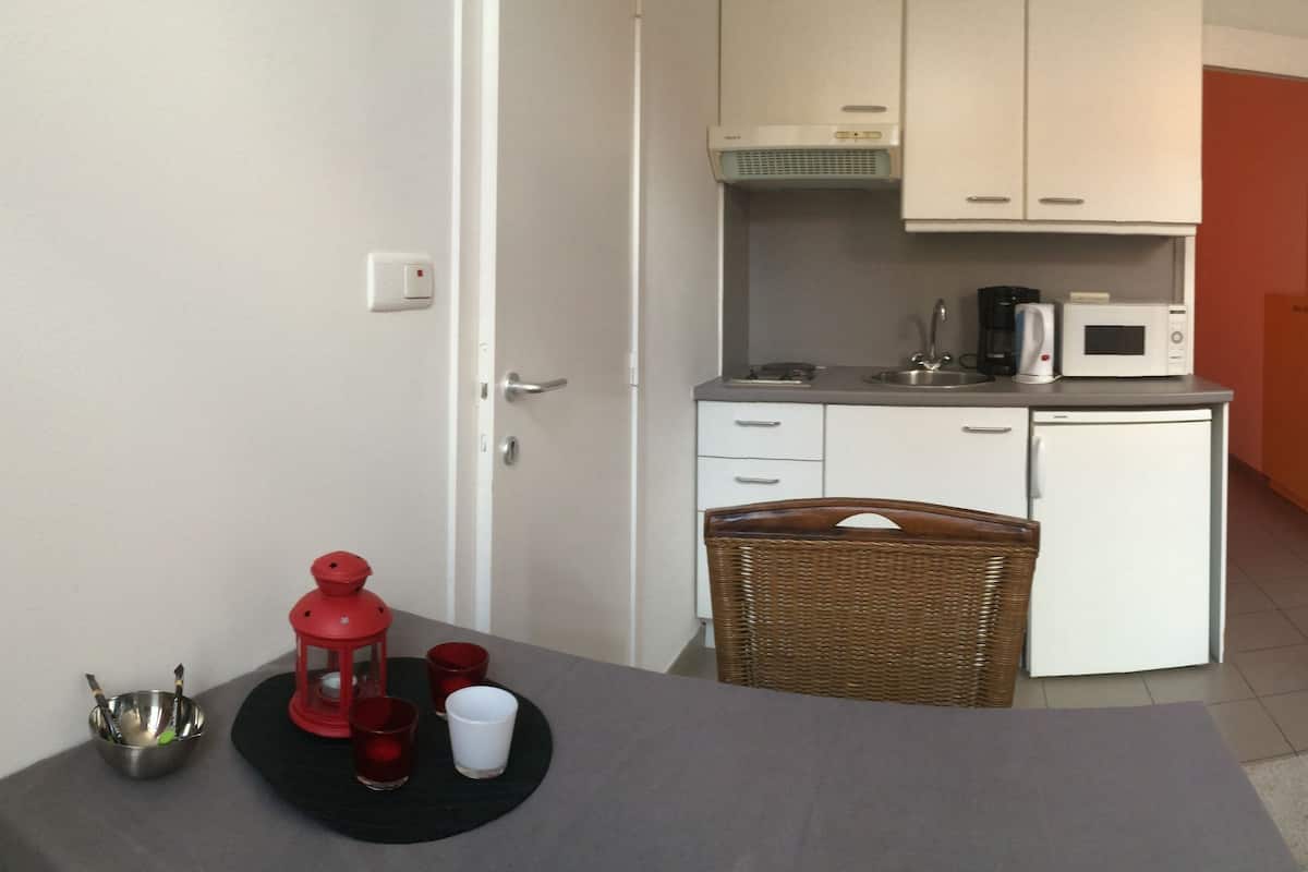 A compact hotel room with a kitchenette, a dining area, and a bed with red bedding.