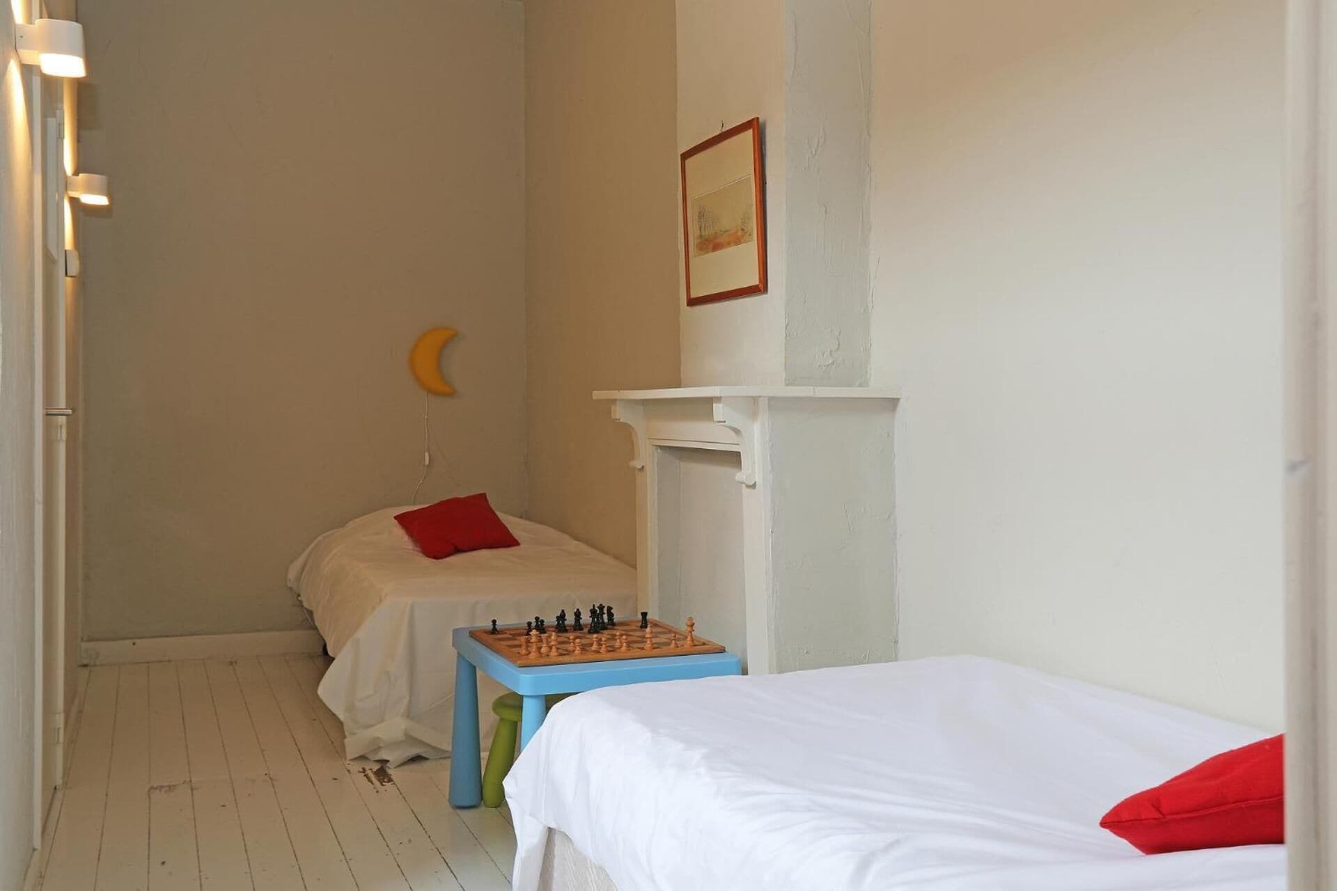 Una habitación pequeña y sencilla con dos camas, una mesita con un tablero de ajedrez y un estante en la pared.