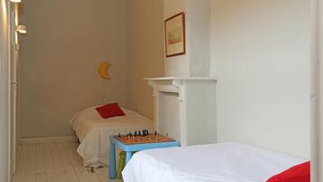 Una habitación pequeña y sencilla con dos camas, una mesita con un tablero de ajedrez y un estante en la pared.