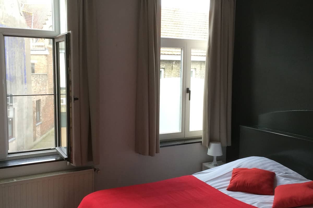 A hotel room with a large bed, red bedding, a window with curtains, a radiator, and a dark headboard.