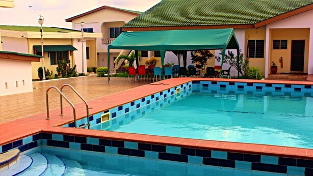 A swimming pool with a tiled edge, a covered seating area with chairs, and a building with a green roof in the background. - Hotel Loreto (Cape Coast)