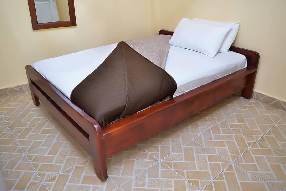 A single bed with a wooden frame, a white mattress, and two pillows, one brown and one white, in a room with tiled flooring.
