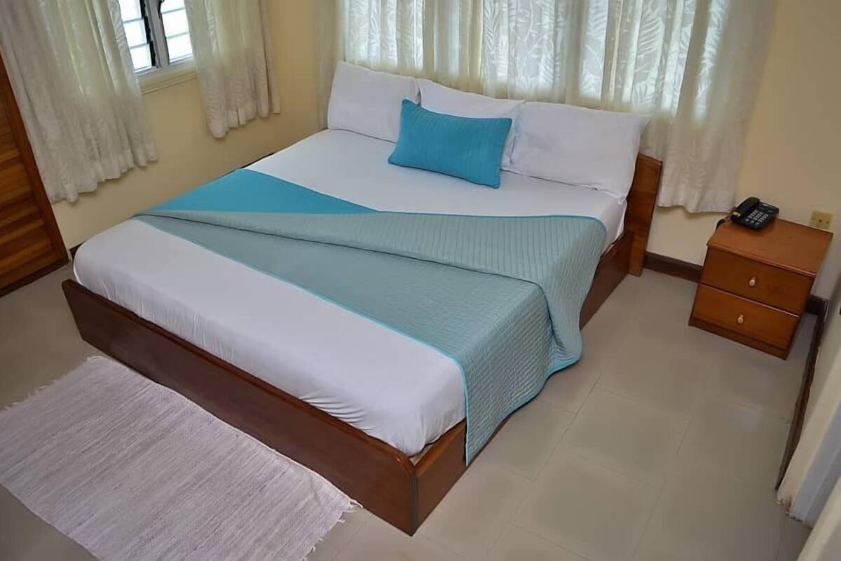 A bedroom with a bed, white curtains, a wooden nightstand, and a white rug.