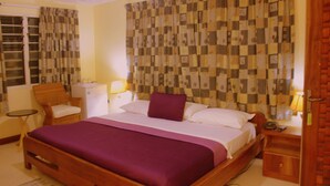 A hotel room with a large bed, a wooden headboard, a small table, a chair, a desk, and a wardrobe. - Hotel Loreto (Cape Coast)