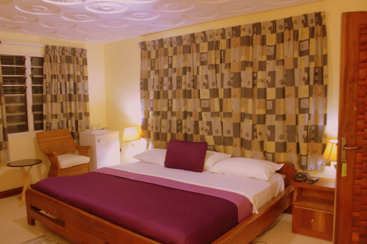 A hotel room with a large bed, a wooden headboard, a small table, a chair, a desk, and a wardrobe.