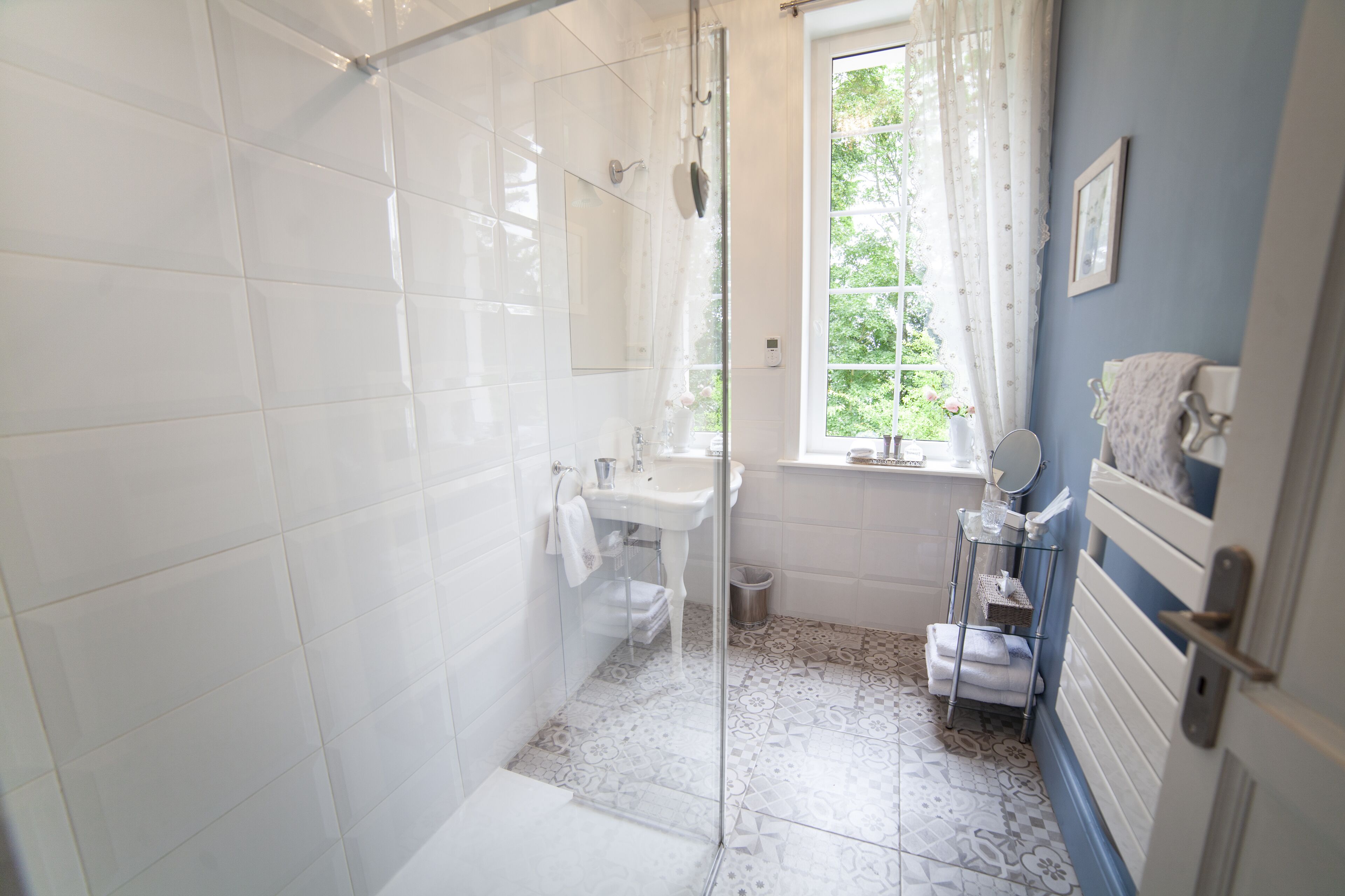 Bagno con piastrelle bianche, box doccia in vetro, lavandino e finestra vista su spazi verdi.