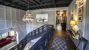 A grand staircase with a dark wooden railing, a chandelier, and a patterned rug leading to a dining area with a table and chairs. - Le Château - Fresnoy en Gohelle (Fresnoy-en-Gohelle)