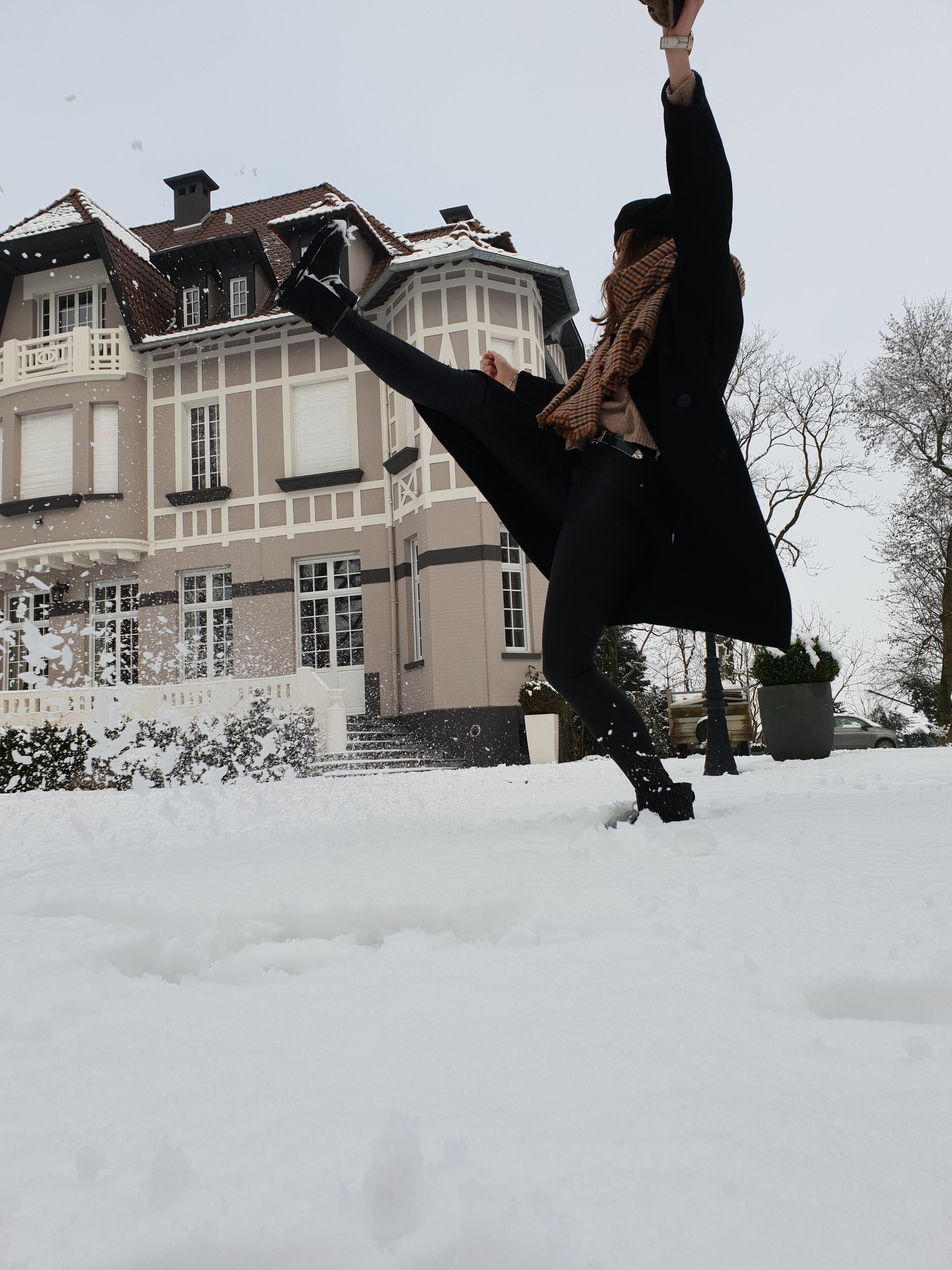 Une personne vêtue de noir joue dans la neige, avec une grande maison en arrière-plan.