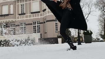Une personne vêtue de noir joue dans la neige, avec une grande maison en arrière-plan.