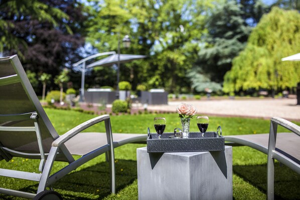 A garden setting with a table set for two, featuring two glasses of red wine and a vase with a single flower.