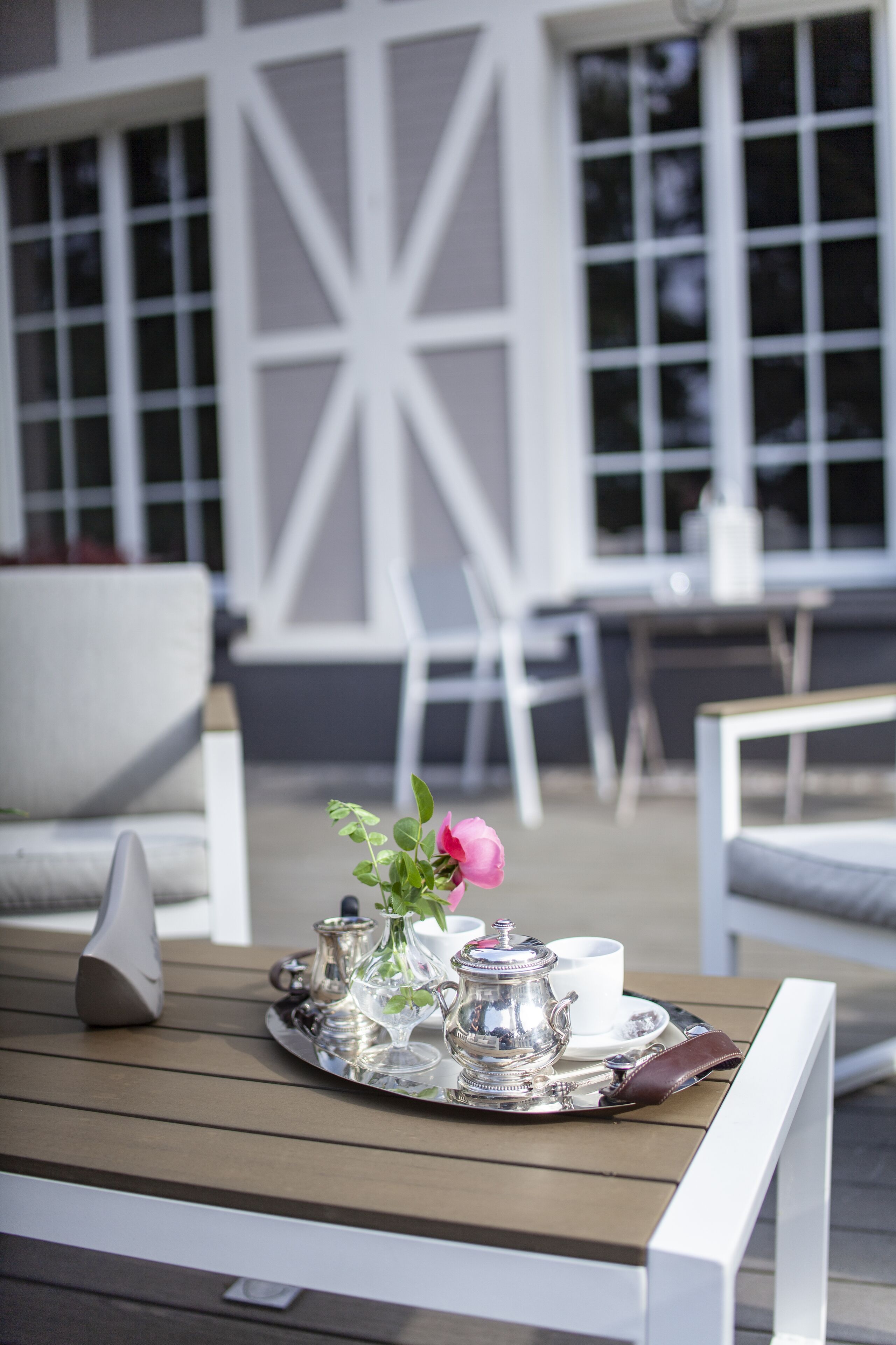 Une table dressée avec un plateau d’argenterie, une théière, des tasses et un vase rempli de fleurs, le tout posé sur une terrasse en bois.