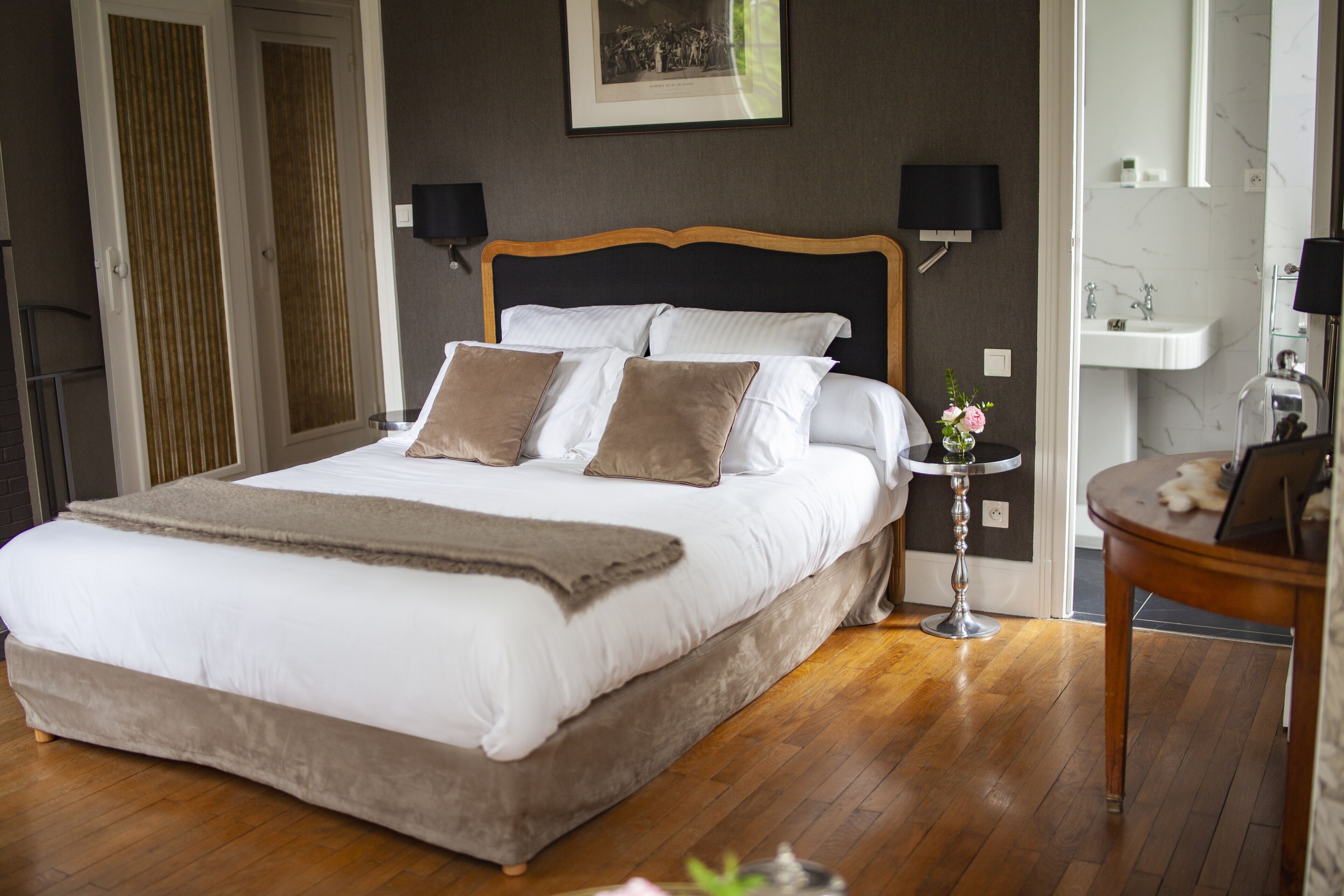 A large bed with a wooden headboard, two bedside tables with lamps, a small round table, and a bathroom with a sink and mirror.