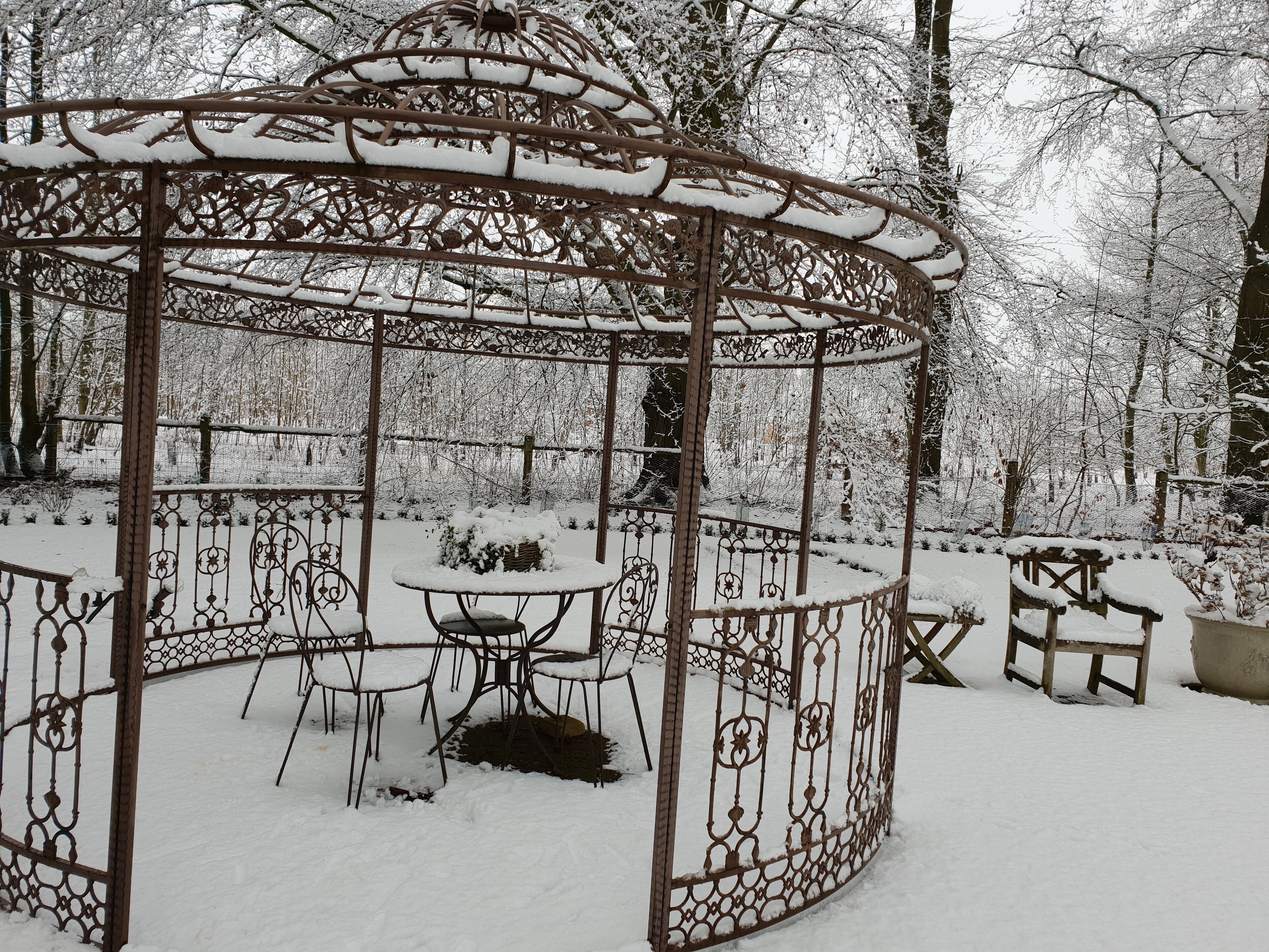 A snow-covered gazebo with a circular table and chairs, surrounded by a metal fence.