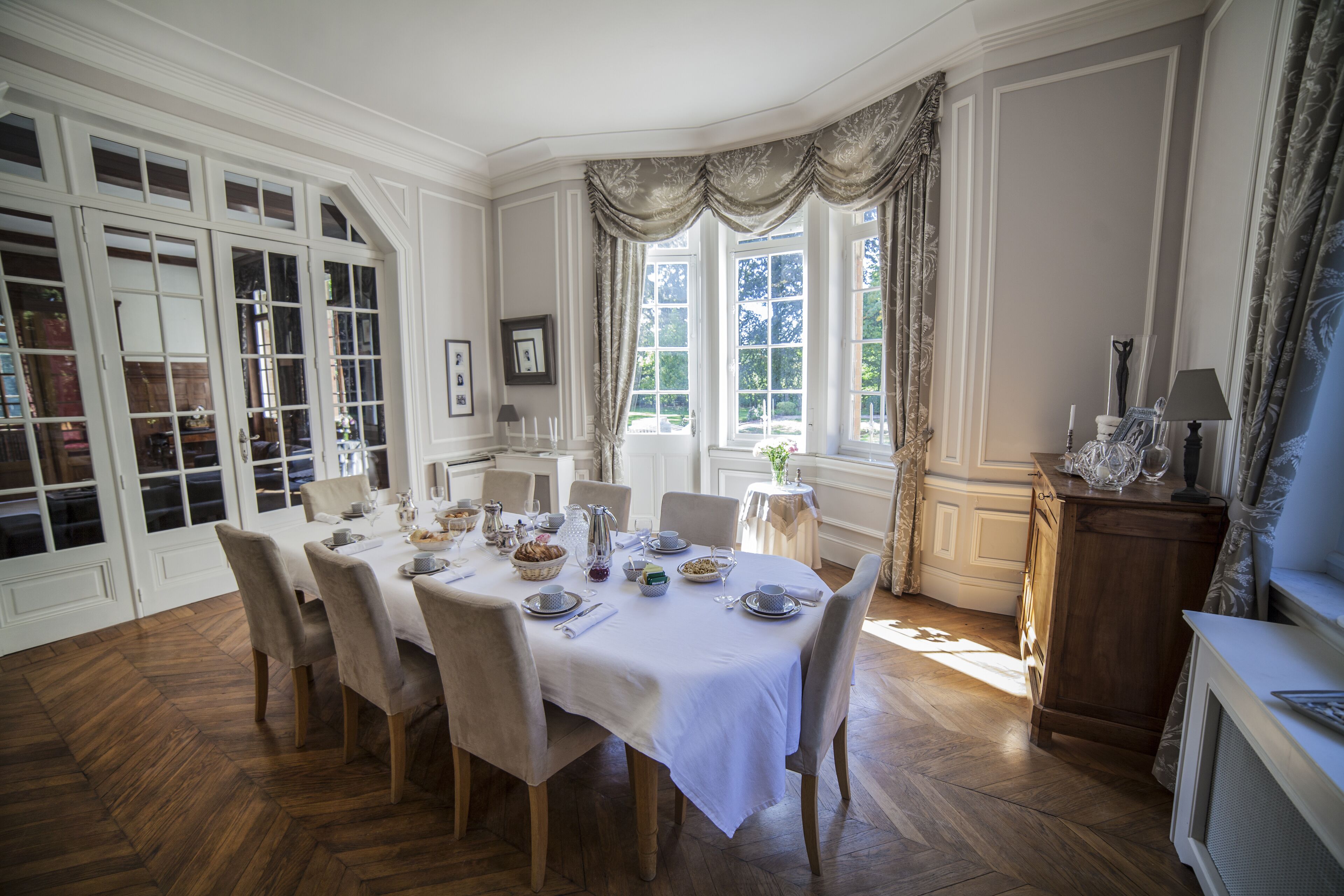 Comedor con una gran mesa dispuesta para una comida, rodeada de sillas. Hay ventanas con cortinas, un aparador de madera y una pequeña mesa con una lámpara.