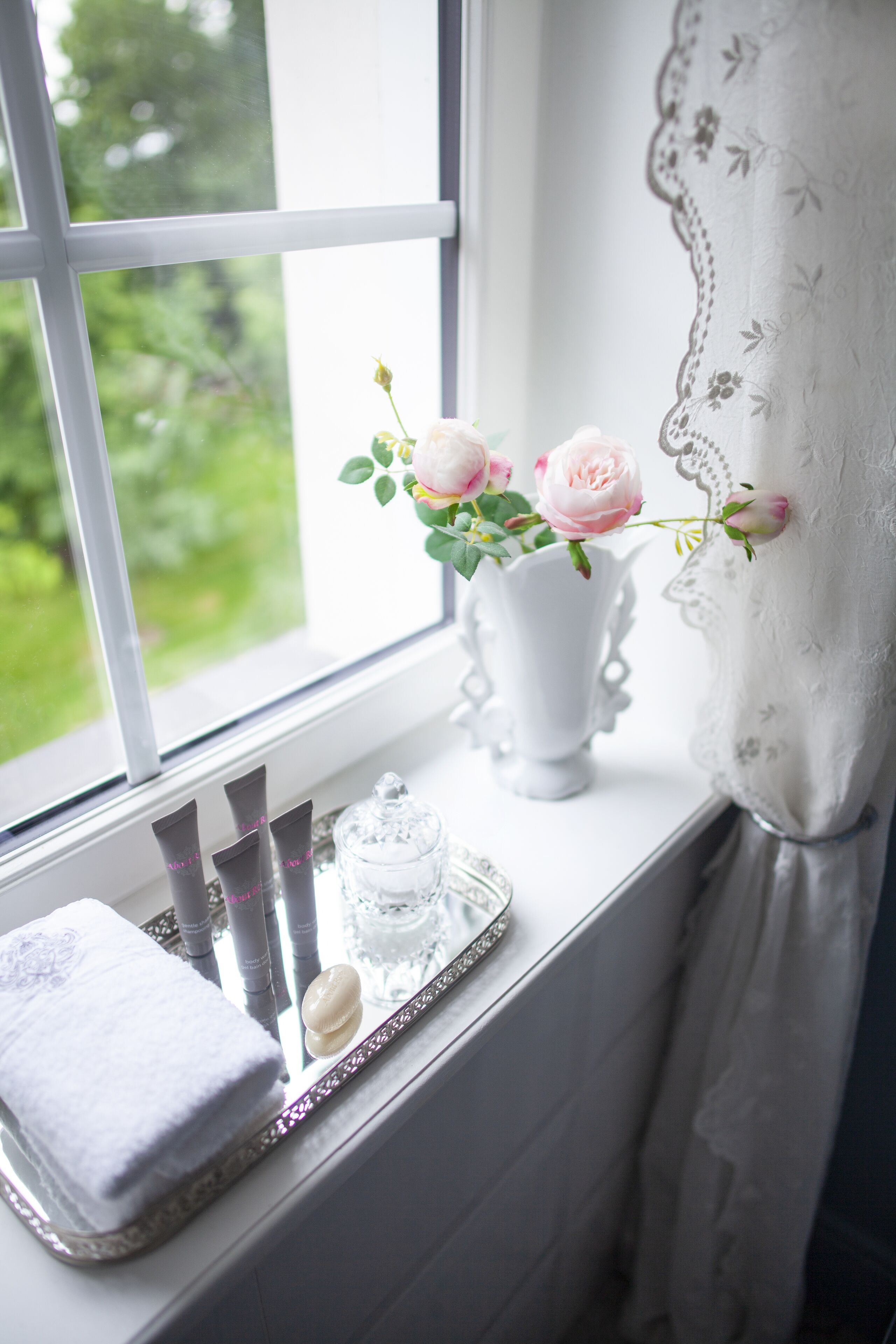 A window with a white vase containing pink roses, a tray with toiletries, and a white towel on a windowsill.