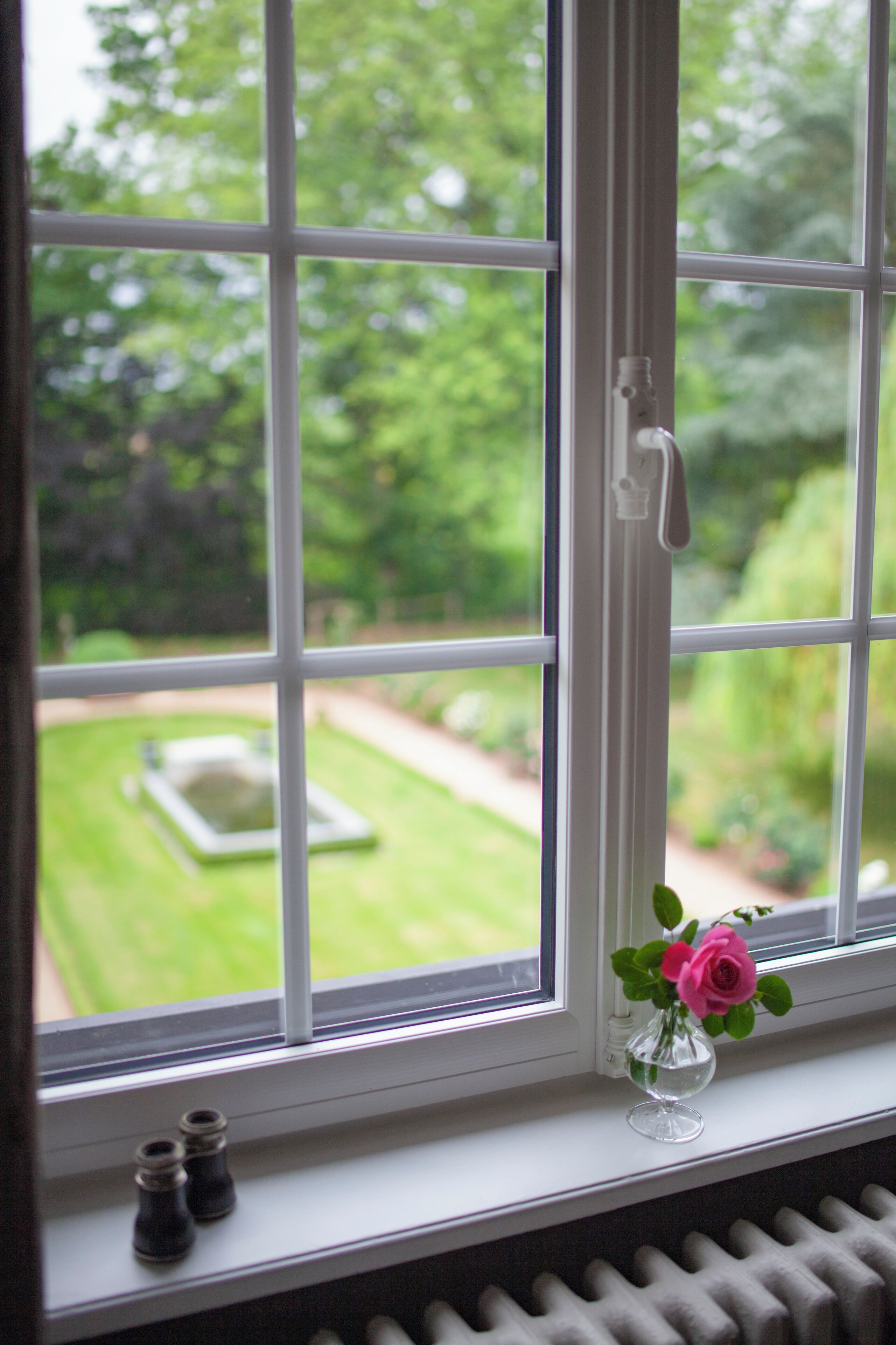 Una ventana con vistas a un jardín, un radiador debajo y un florero con una flor en el alféizar.