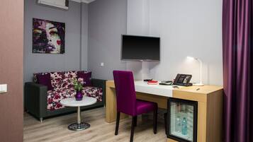 Una habitación moderna con sofá, escritorio, silla, televisor, una mesita con un jarrón y un mini refrigerador.