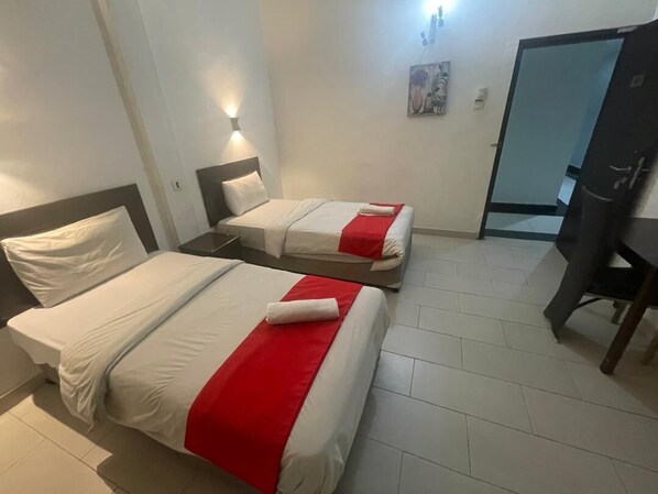 Habitación de hotel con dos camas, cada una con ropa blanca y detalles rojos, una mesita de noche, un escritorio y una puerta.
