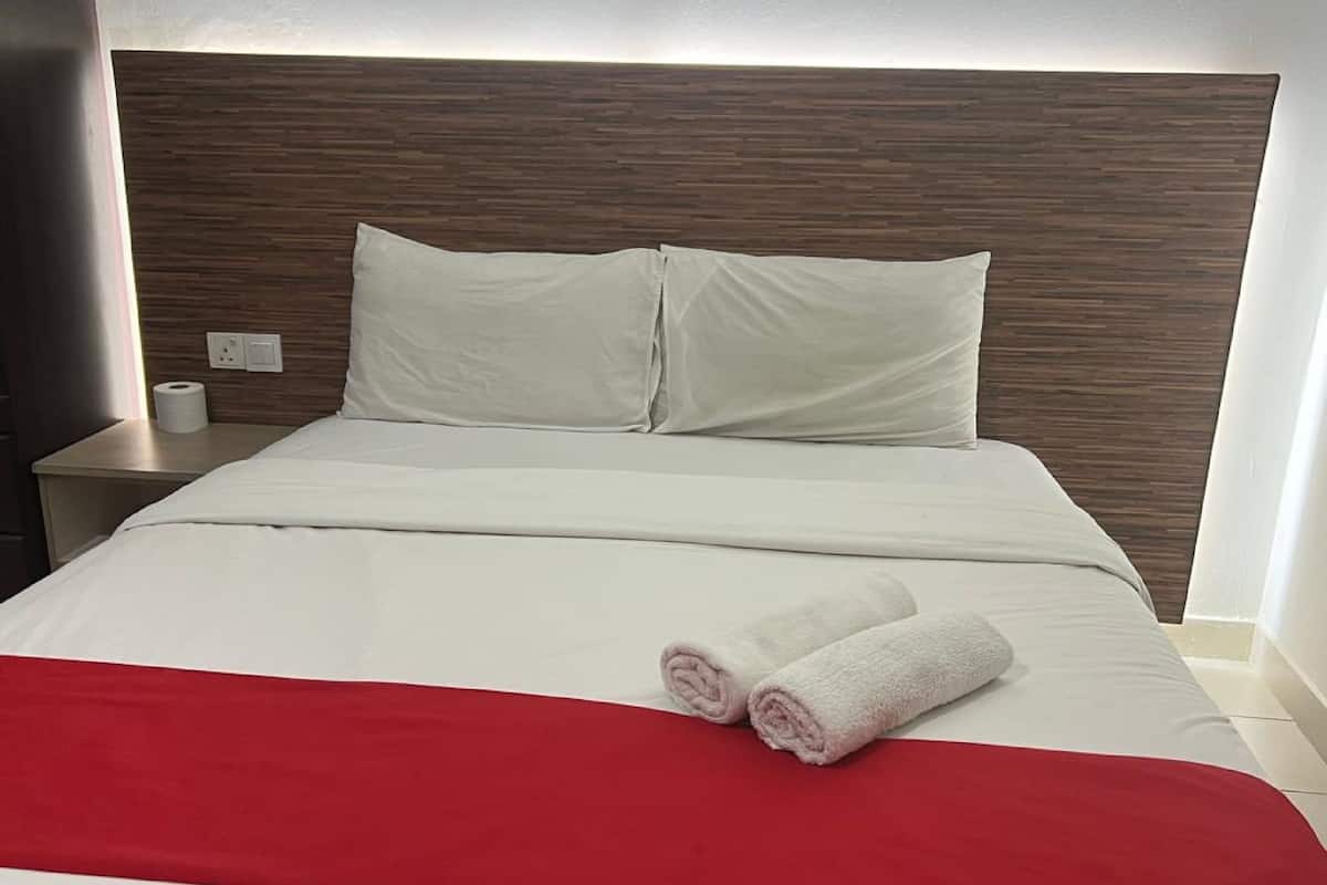 A neatly made bed with white linens and a red blanket, two pillows, and two rolled towels on the bed.