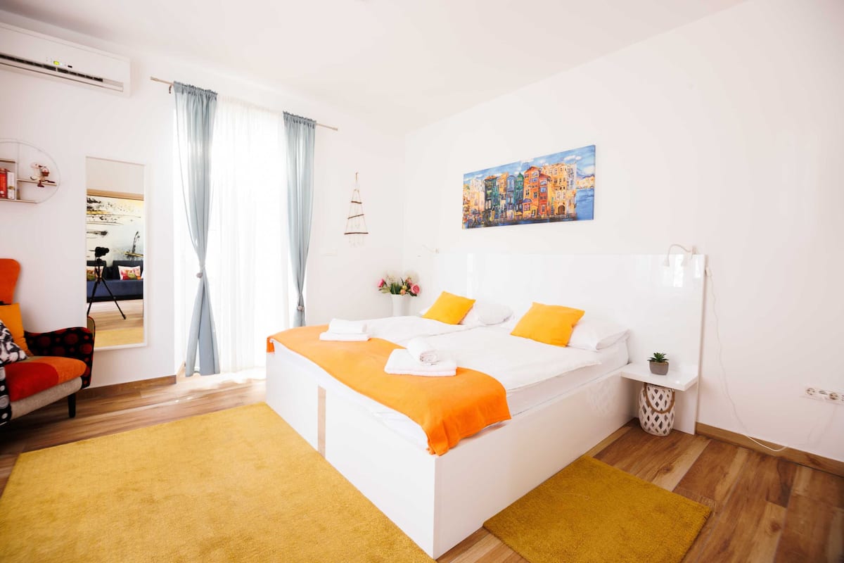 Una habitación de hotel moderna con una cama grande, una mesita auxiliar pequeña, un cuadro en la pared y una ventana con cortinas.