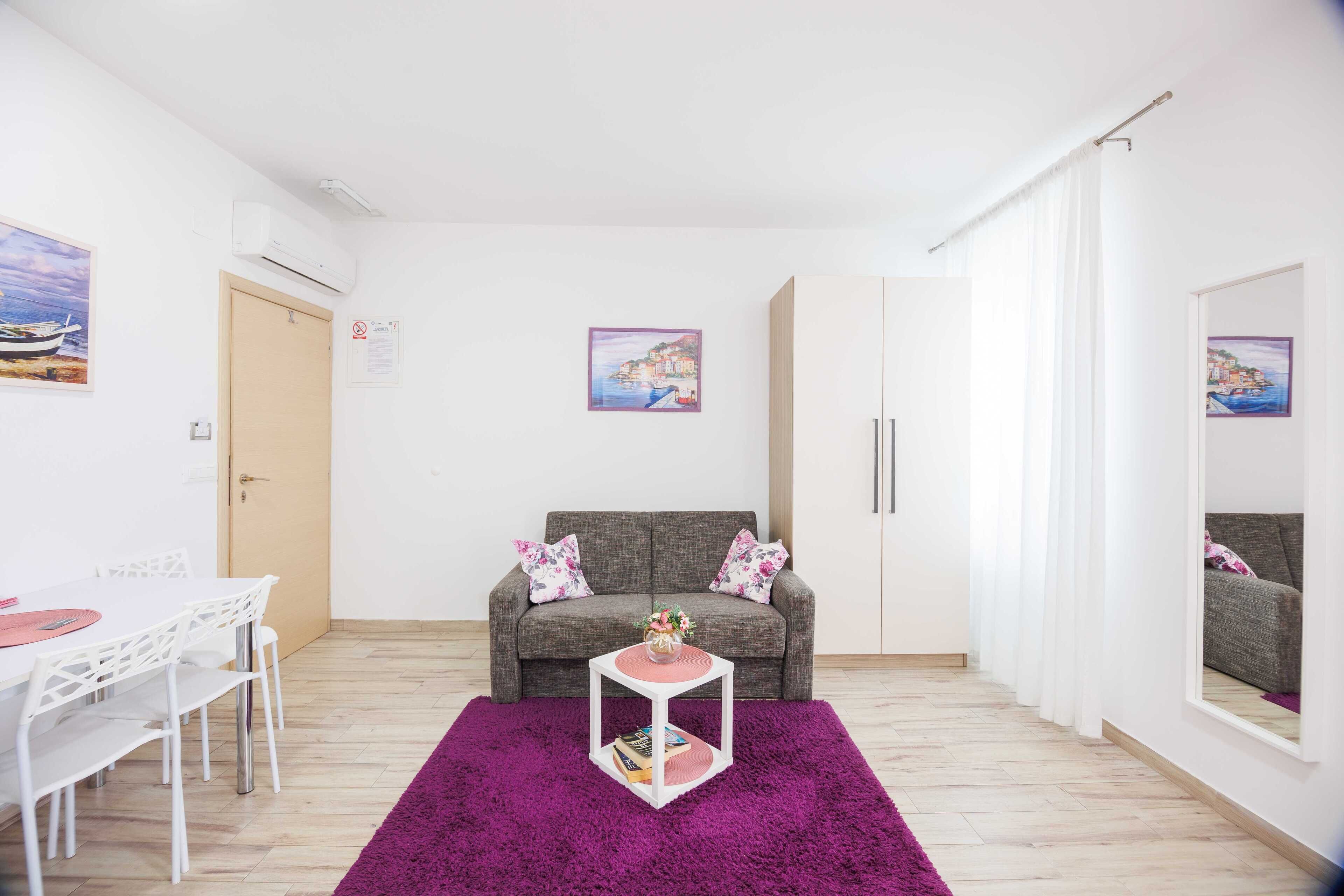 Una sala moderna con un sofá gris, una mesa de centro blanca y una alfombra púrpura. Hay una mesa de comedor con sillas y un cuadro en la pared.