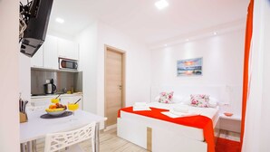 Une chambre d’hôtel compacte avec une kitchenette, un lit recouvert d’une couverture rouge, un tableau au mur et un tapis rouge.