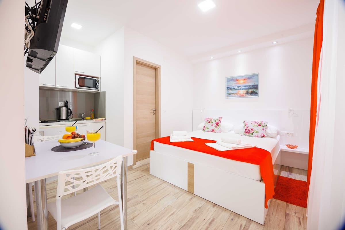 Una habitación de hotel compacta con kitchenette, una cama con manta roja, un cuadro en la pared y una alfombra roja.