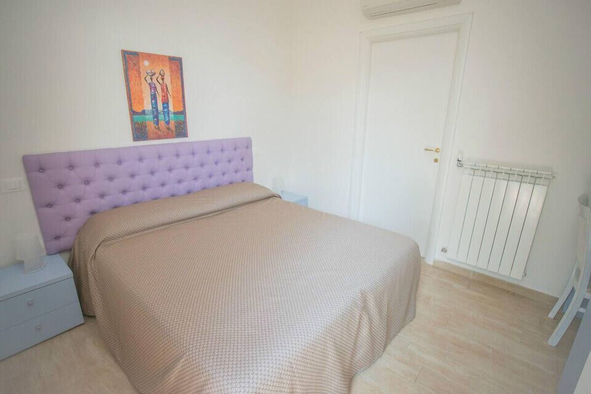 Ein Schlafzimmer mit violettem, gesteptem Kopfteil, einer beigefarbenen Bettdecke, einem Nachttisch, einem Heizkörper und einer Tür.