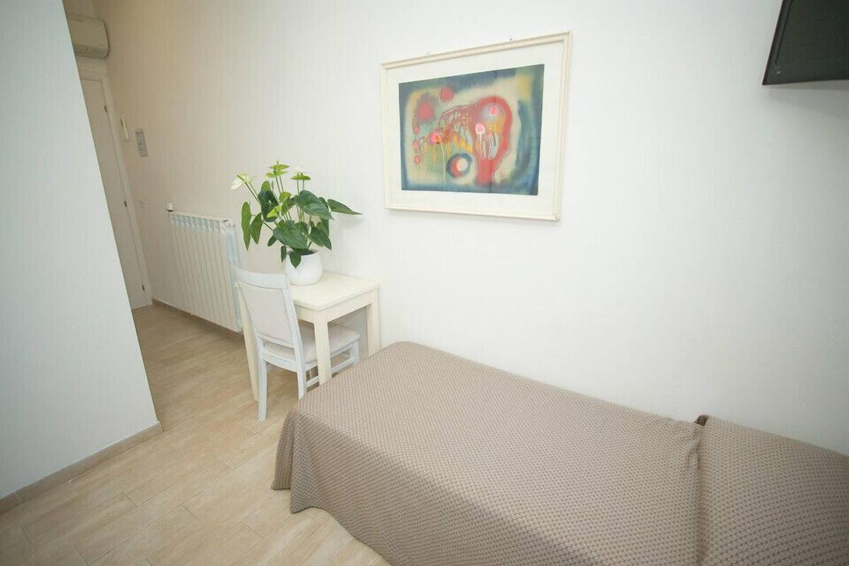 Ein kleiner, ordentlicher Raum mit einem Bett, einem weißen Stuhl, einem kleinen Tisch, einer Topfpflanze und einem gerahmten abstrakten Gemälde an der Wand.