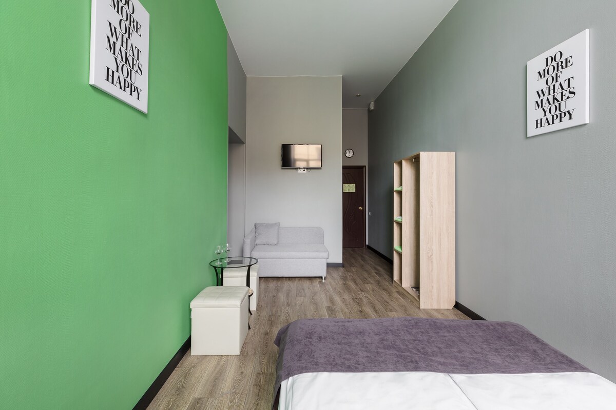 A modern hotel room with a green accent wall, a grey sofa, a small white table, a bed with a purple blanket, and two motivational posters on the walls.
