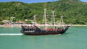 Un gran barco velero con varios mástiles y pasajeros, navegando cerca de una playa con colinas verdes y exuberantes al fondo.