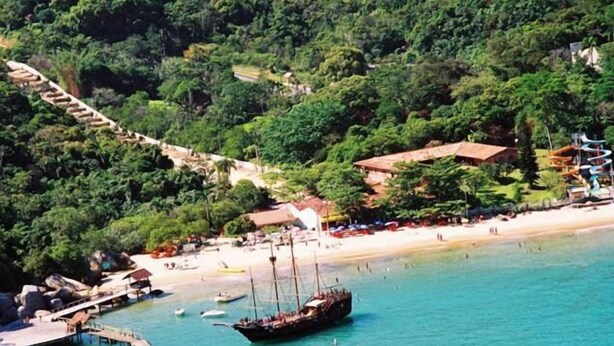 Zona costera con barcos amarrados en un muelle, una playa y una colina con edificios.