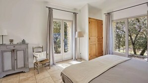 Um quarto com cama, porta de madeira, cadeira, armário e janelas amplas com cortinas.