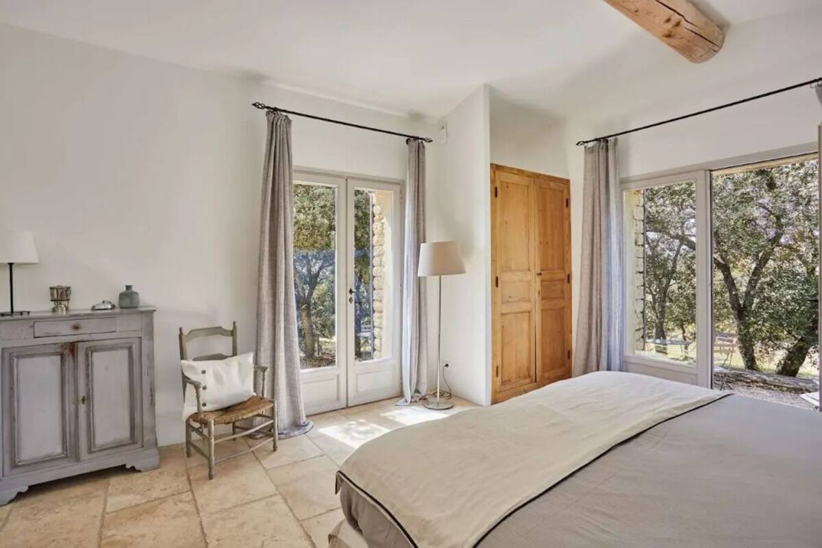 A bedroom with a bed, a wooden door, a chair, a cabinet, and large windows with curtains.