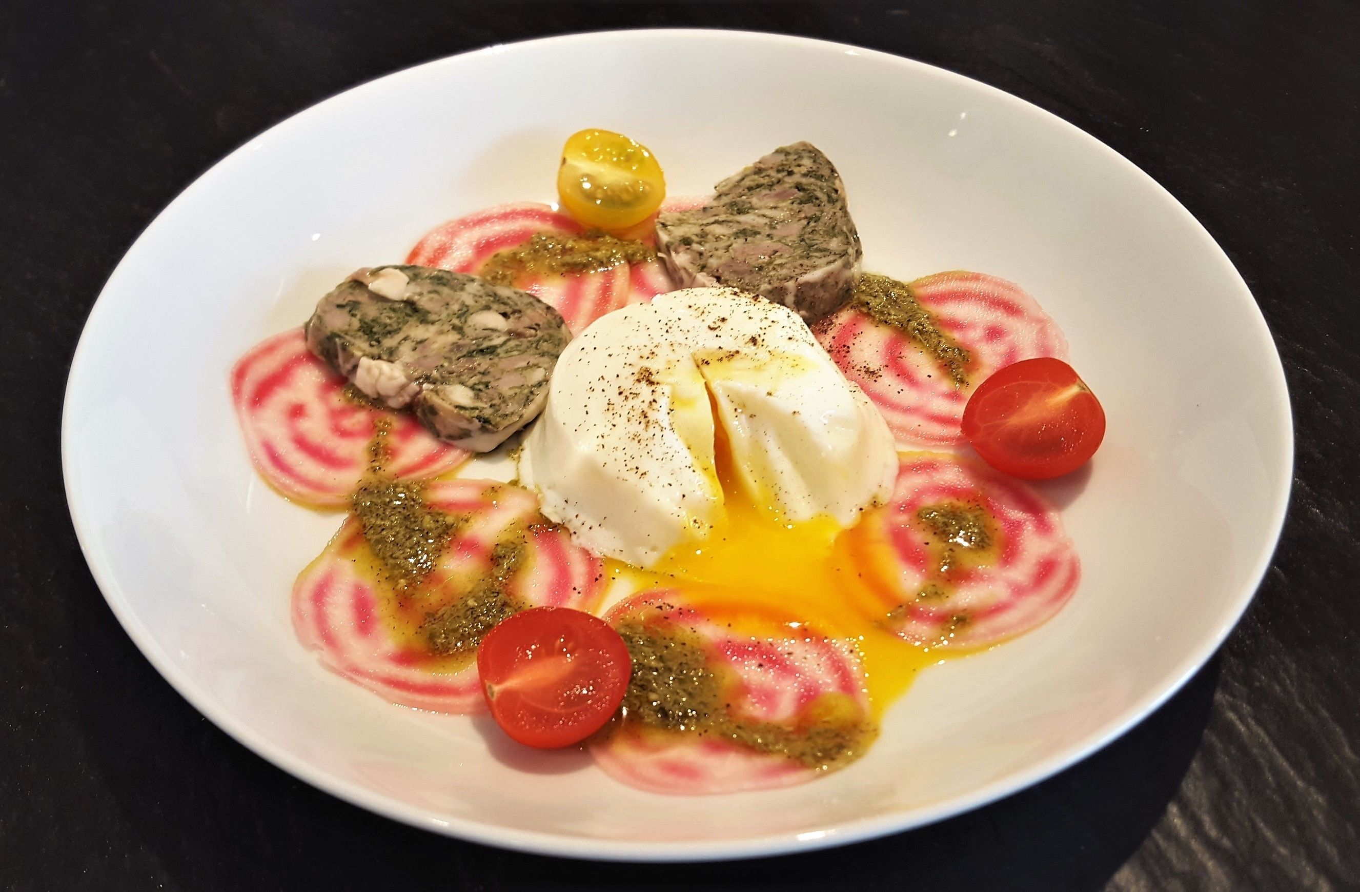 A white plate with a dish containing a poached egg, cherry tomatoes, and a green sauce.